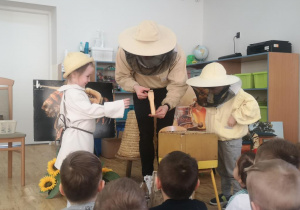 Widok na pszczelarza i dzieci prezentujące akcesoria pracy pszczelarza.
